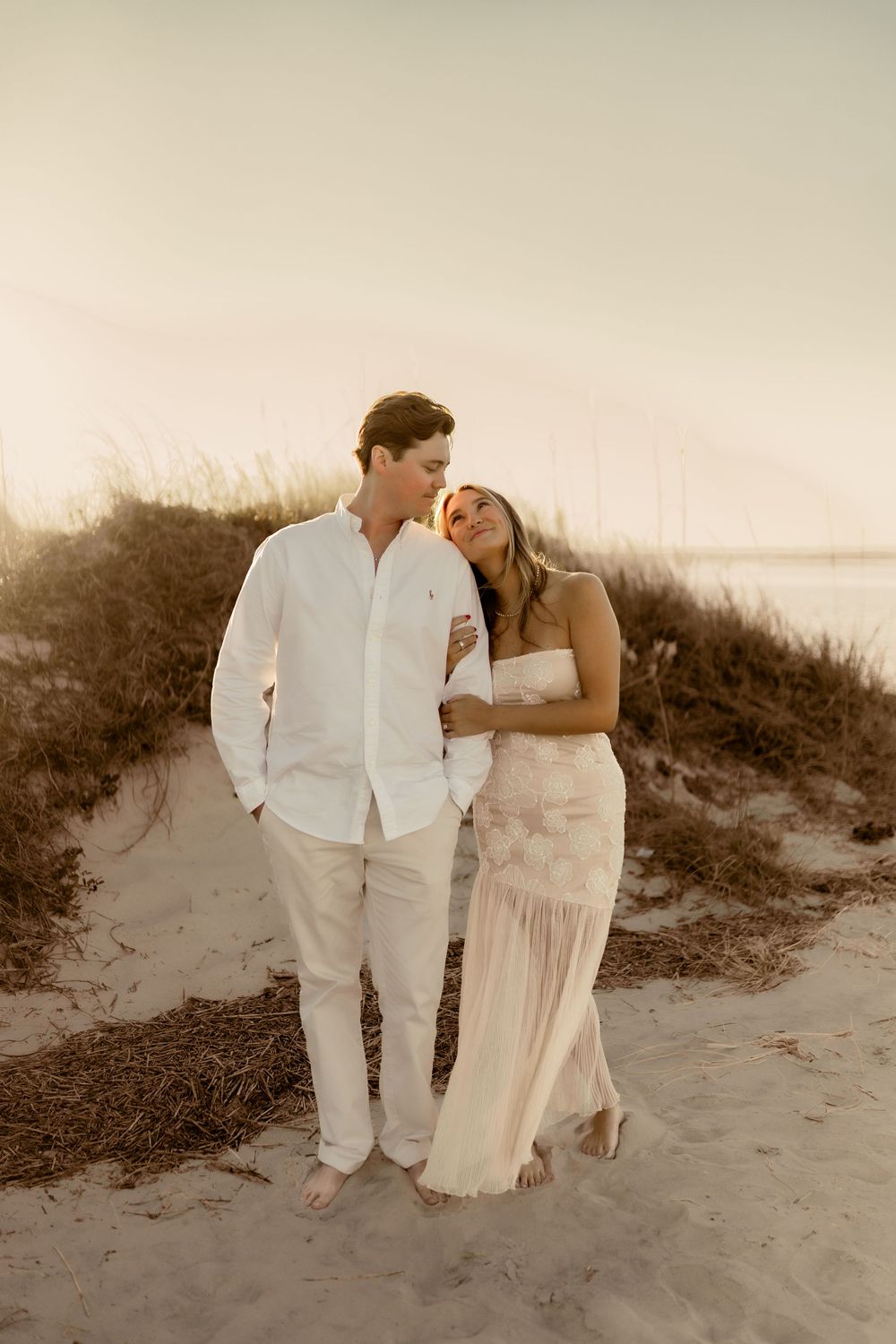 Romantic couple in white attire share intimate moments on a sandy beach at sunset.