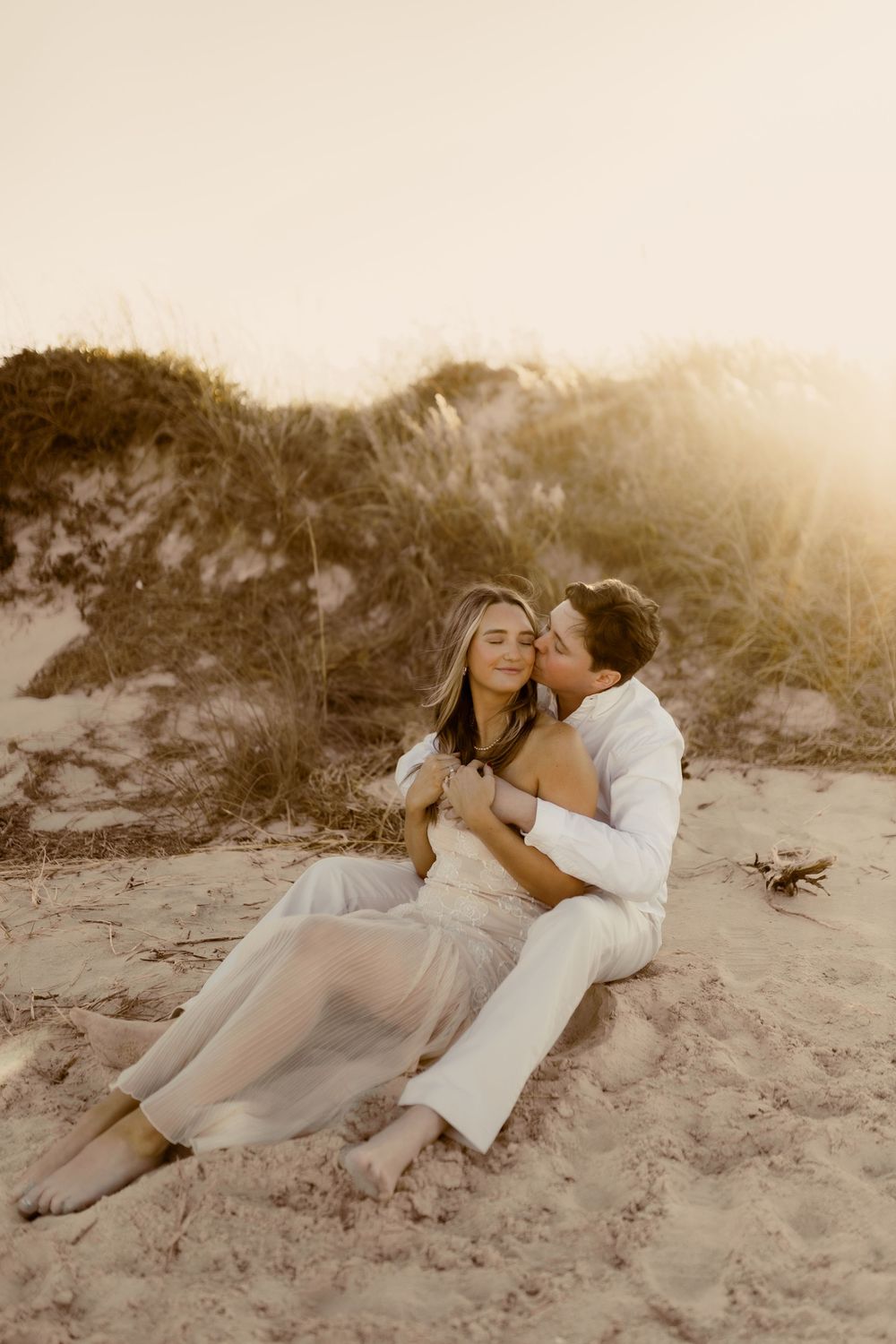 A romantic couple embraces on a sandy beach at sunset with sand dunes in the background.