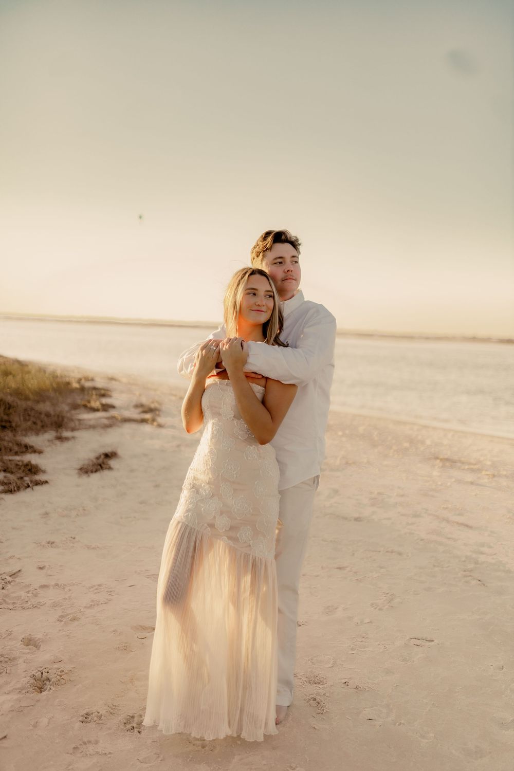 A couple in white outfits embracing on a sandy beach during golden hour.