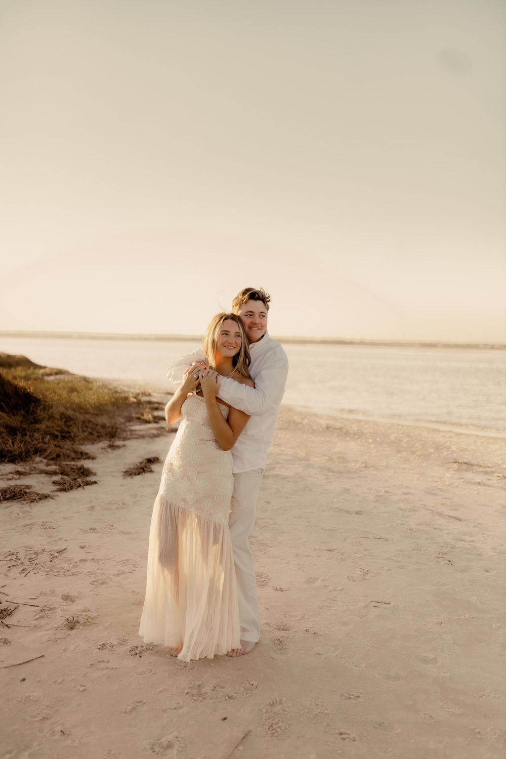A romantic beach photoshoot featuring a couple in white outfits embracing by the ocean at golden hour.