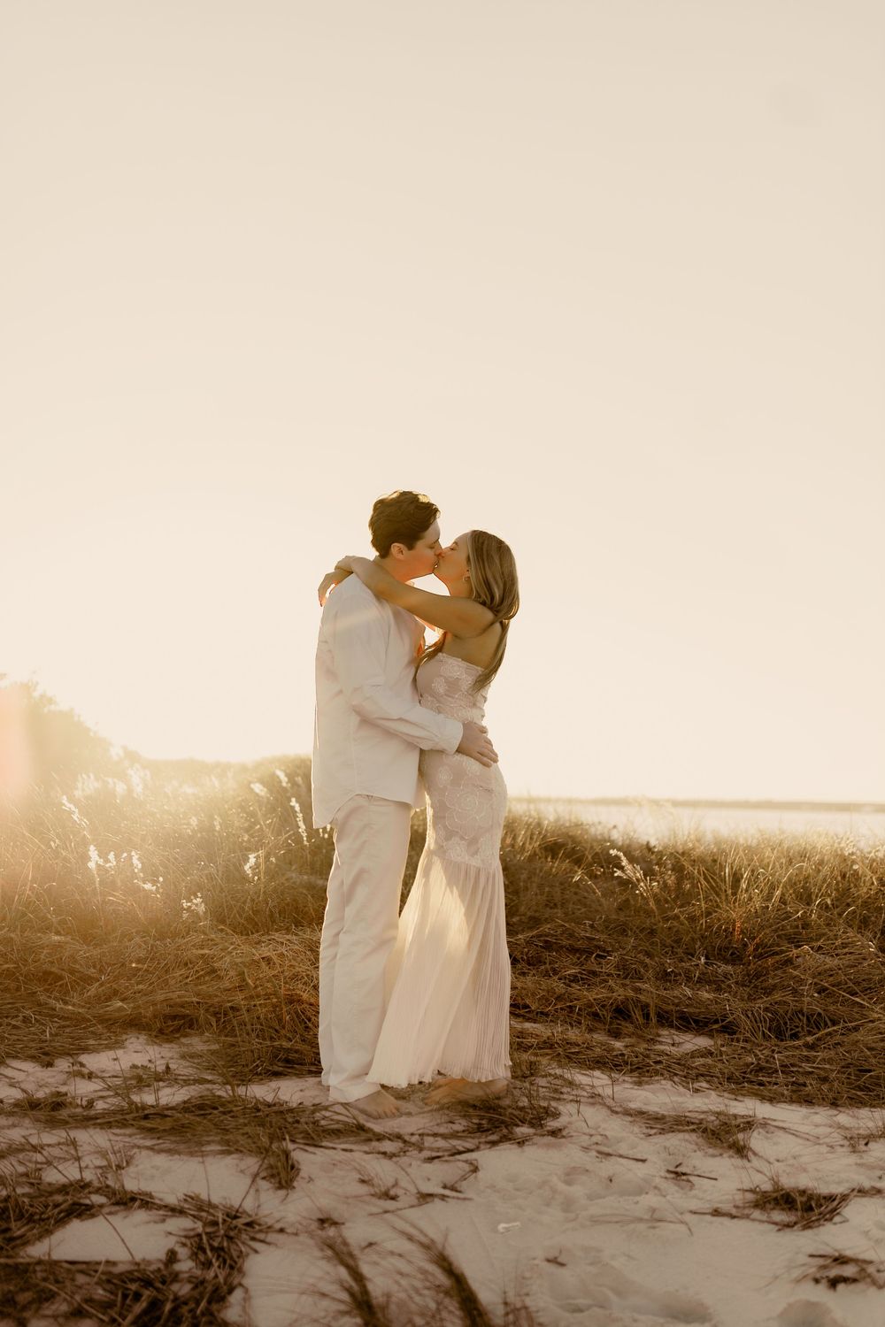 Romantic sunset beach photo of couple in white embracing on sandy dunes.