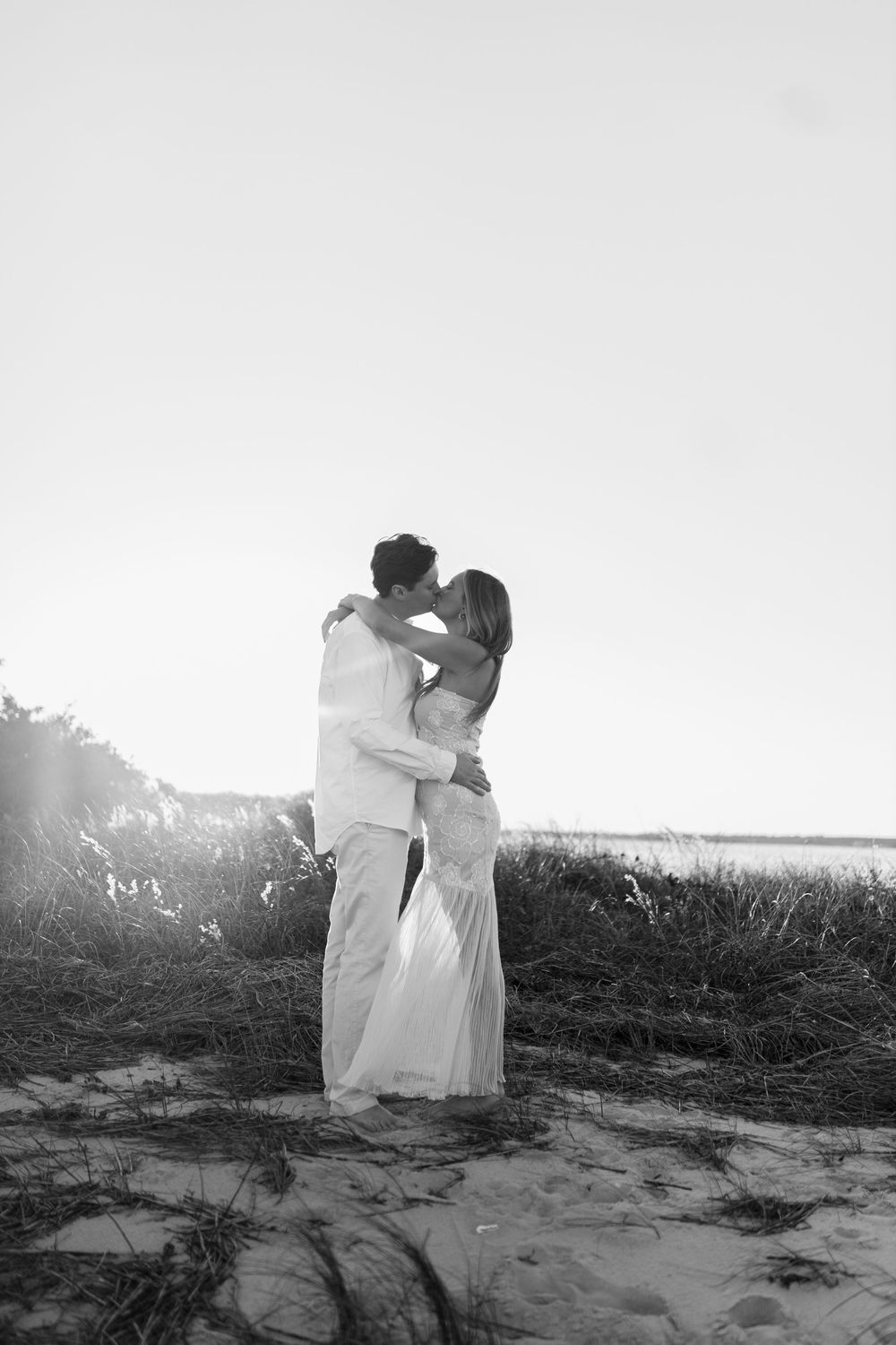 A black and white photograph captures an intimate moment between a couple on a coastal dune.