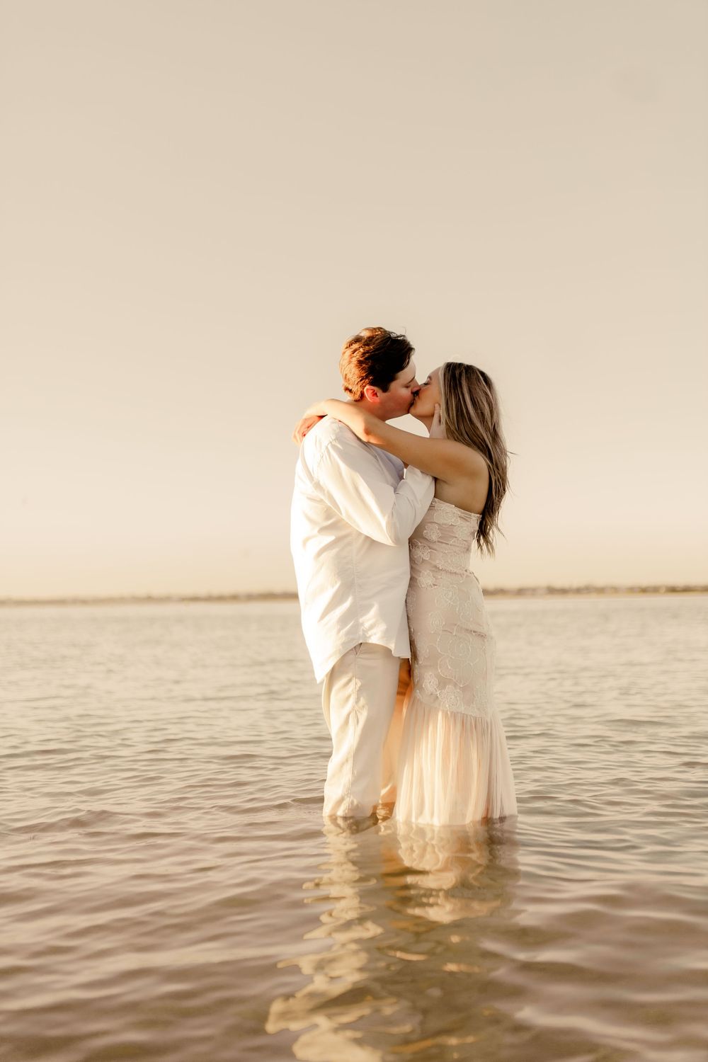 A romantic couple standing in shallow ocean water during sunset, both wearing white.