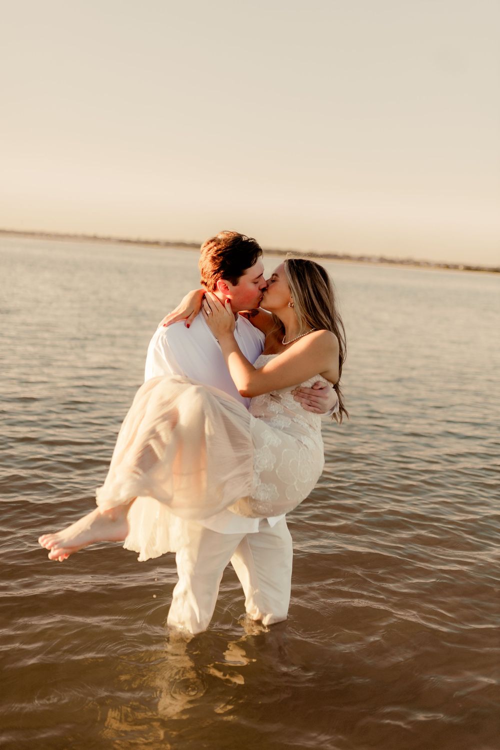 A couple in white wedding attire shares a romantic kiss while standing in shallow ocean water at sunset.