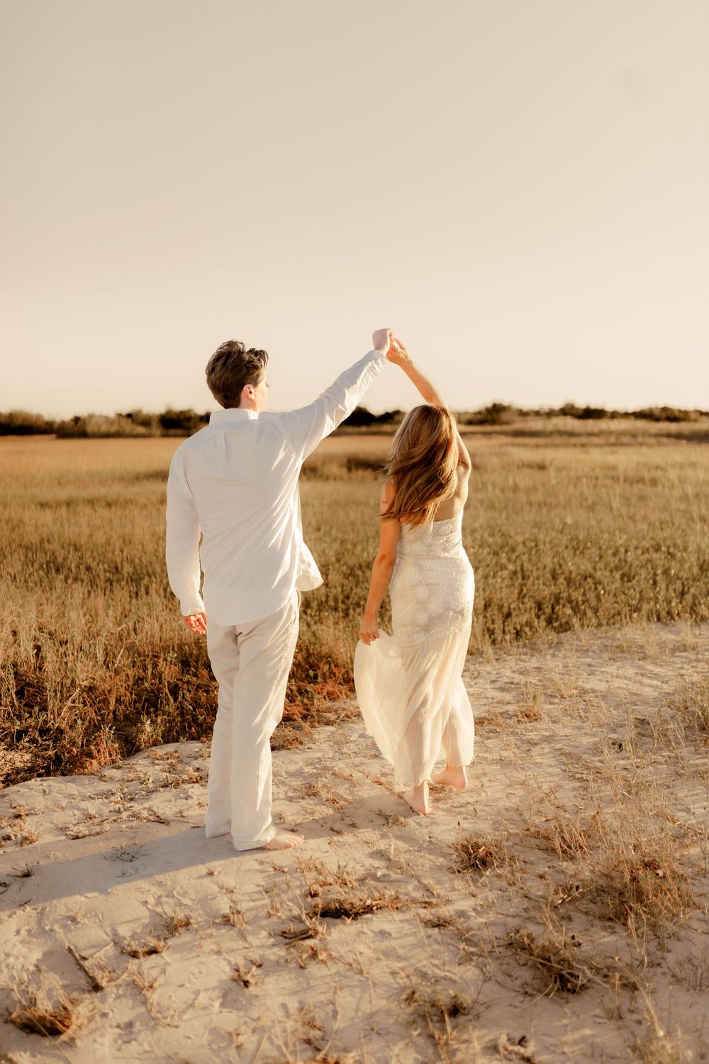 A couple in white outfits walks hand in hand through a golden field at sunset.