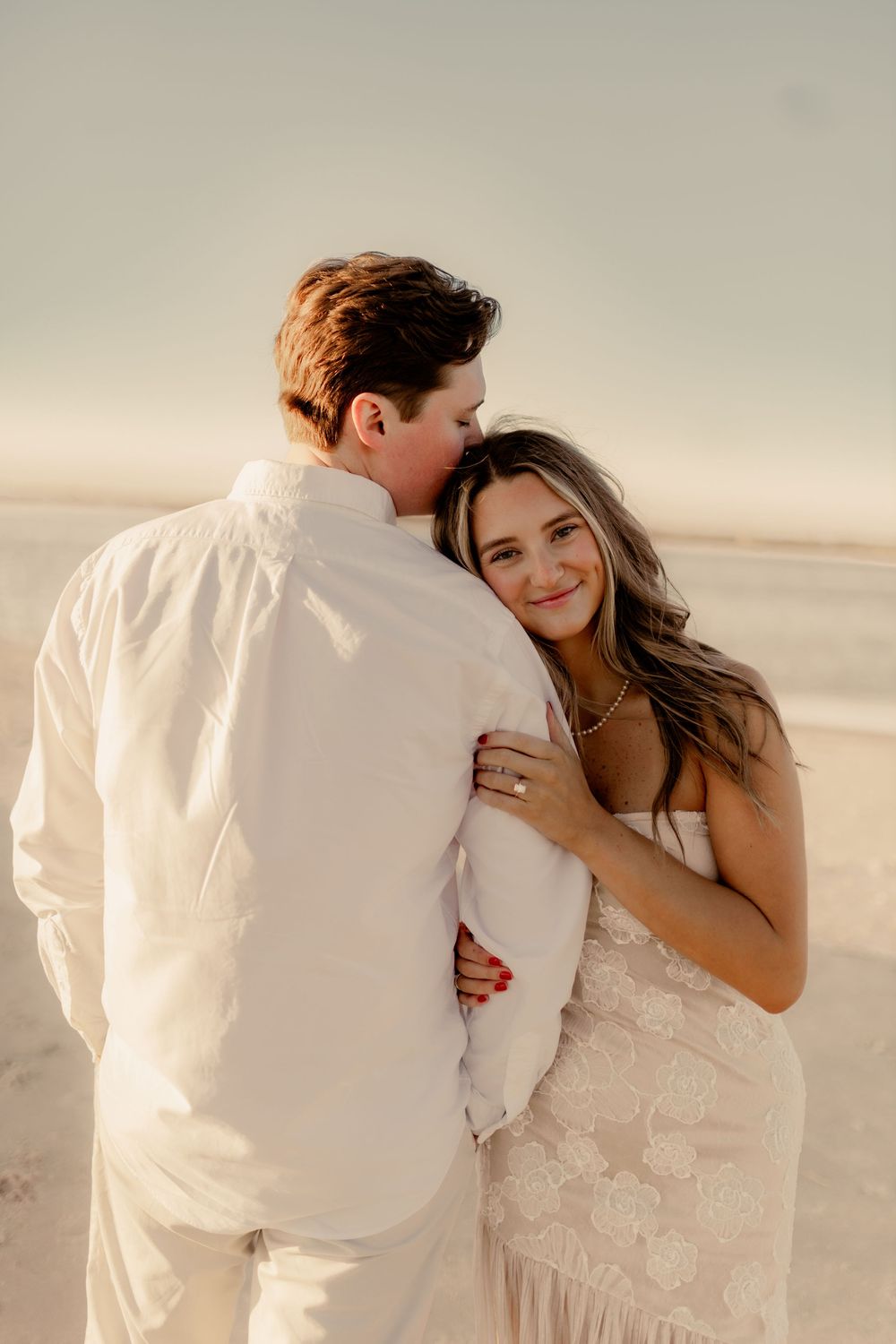 A tender moment between a couple in white attire during their beach engagement photoshoot at sunset.