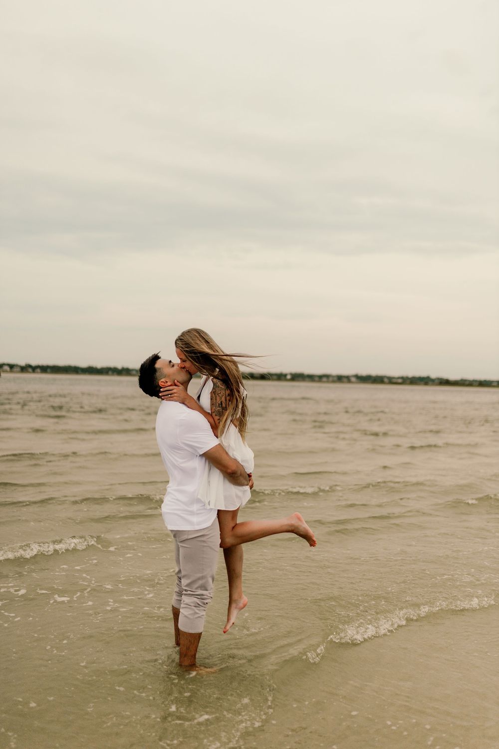 A couple in white outfits shares a romantic moment on the beach, as the man lifts the woman near the ocean waves.