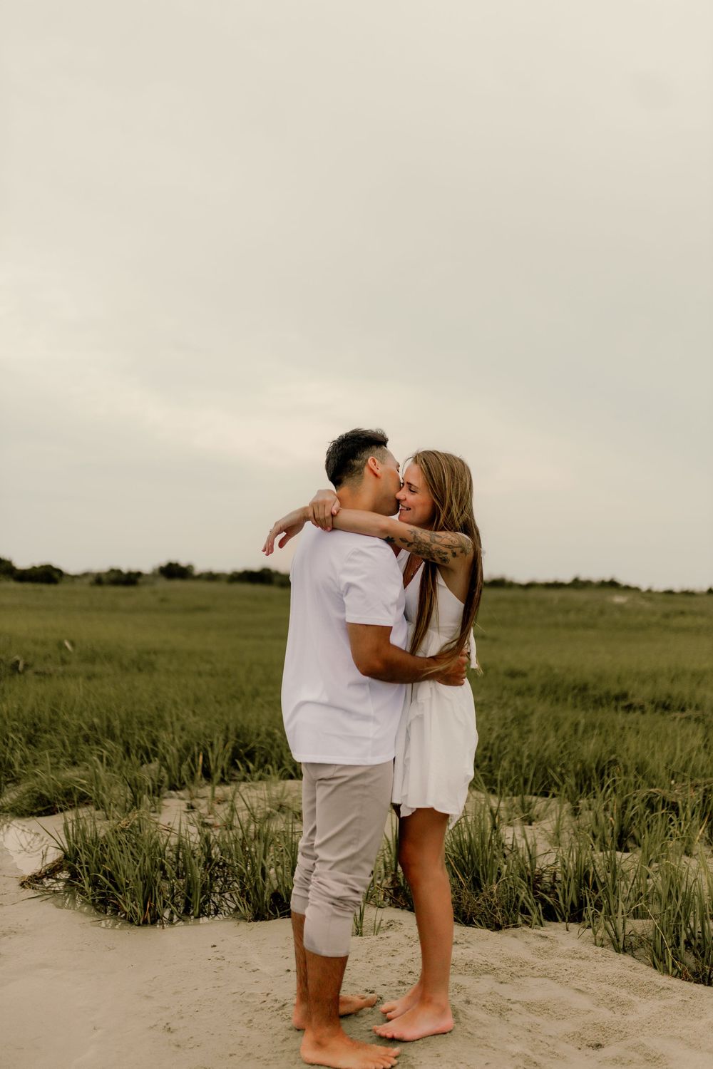 Couple embracing and kissing on sandy beach path surrounded by green marsh grass in matching white outfits.