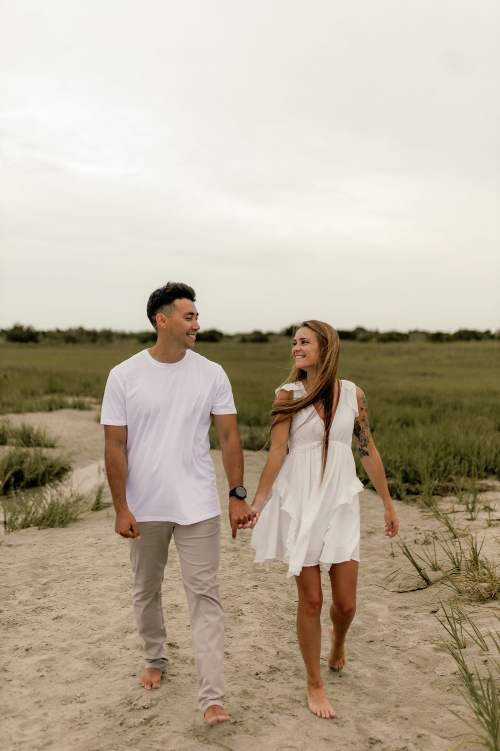 Couple holding hands walking on sandy beach path, woman in white dress, man in white tee, green marsh background.