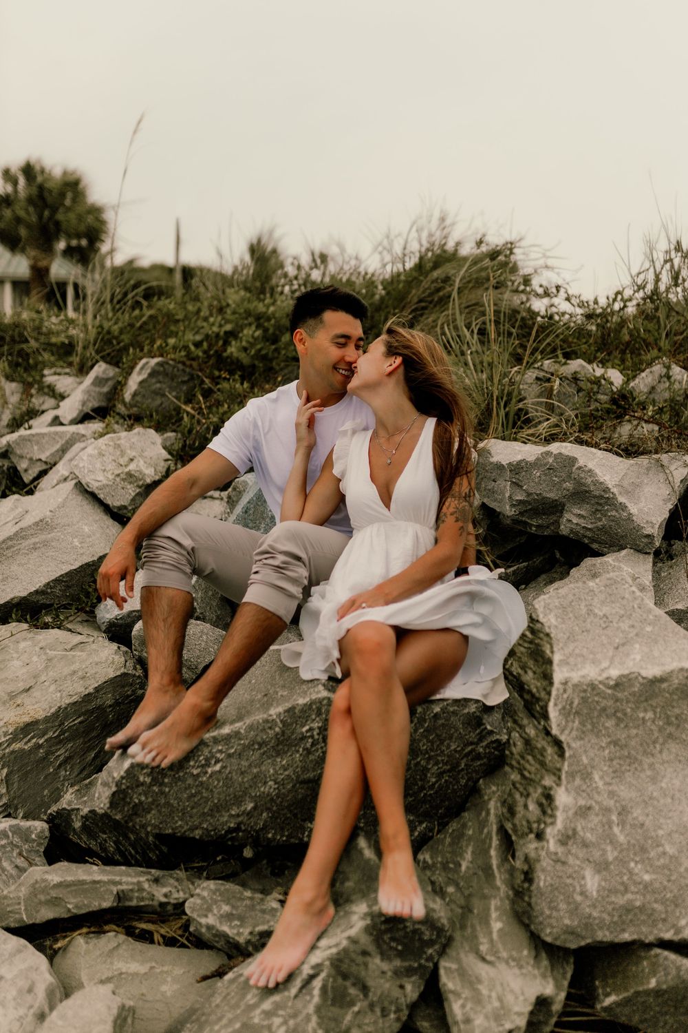Romantic couple sitting barefoot on rocks near the beach, sharing an intimate moment in casual summer outfits.