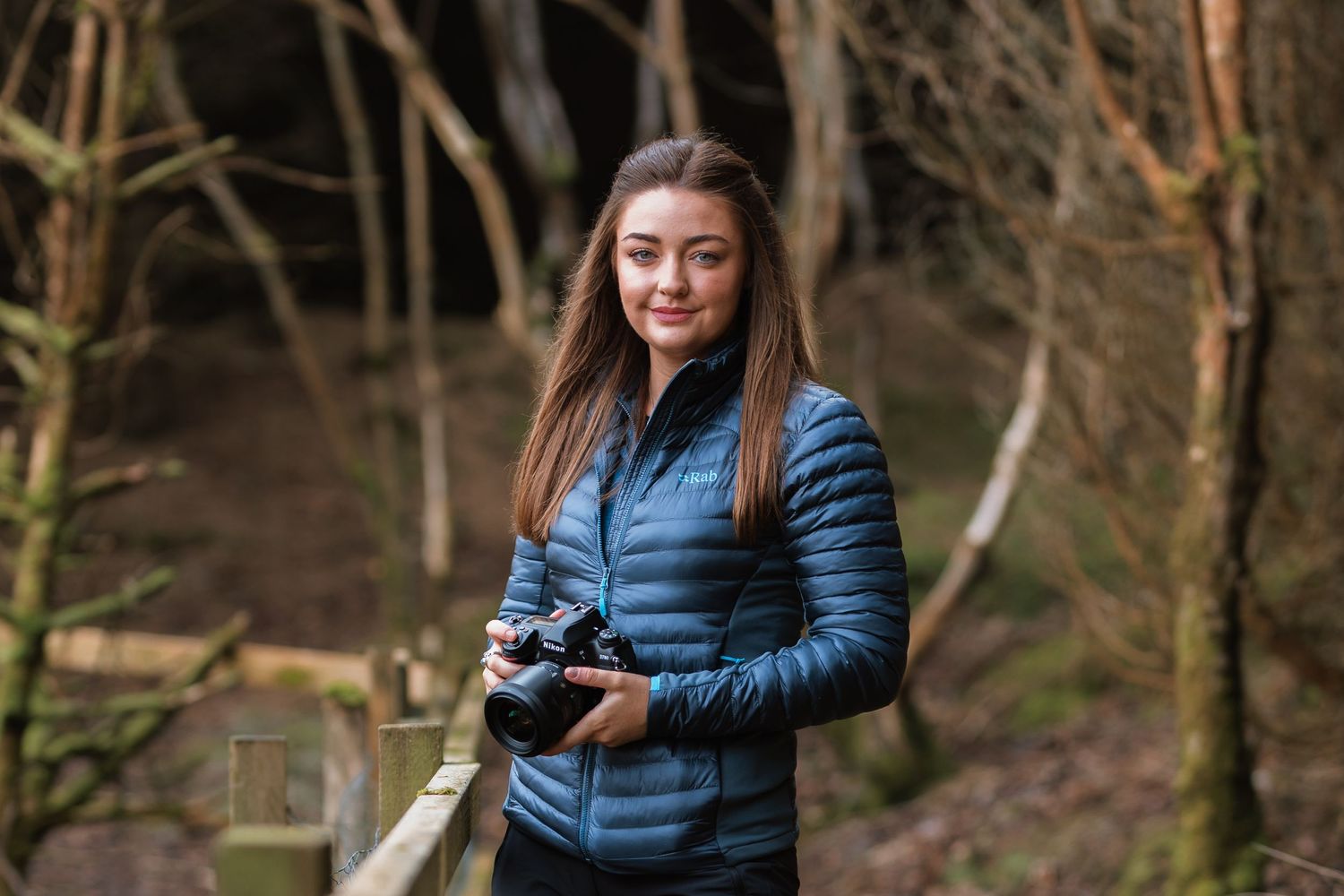 Photographer in blue puffer jacket holding camera while exploring forest trail on winter day.
