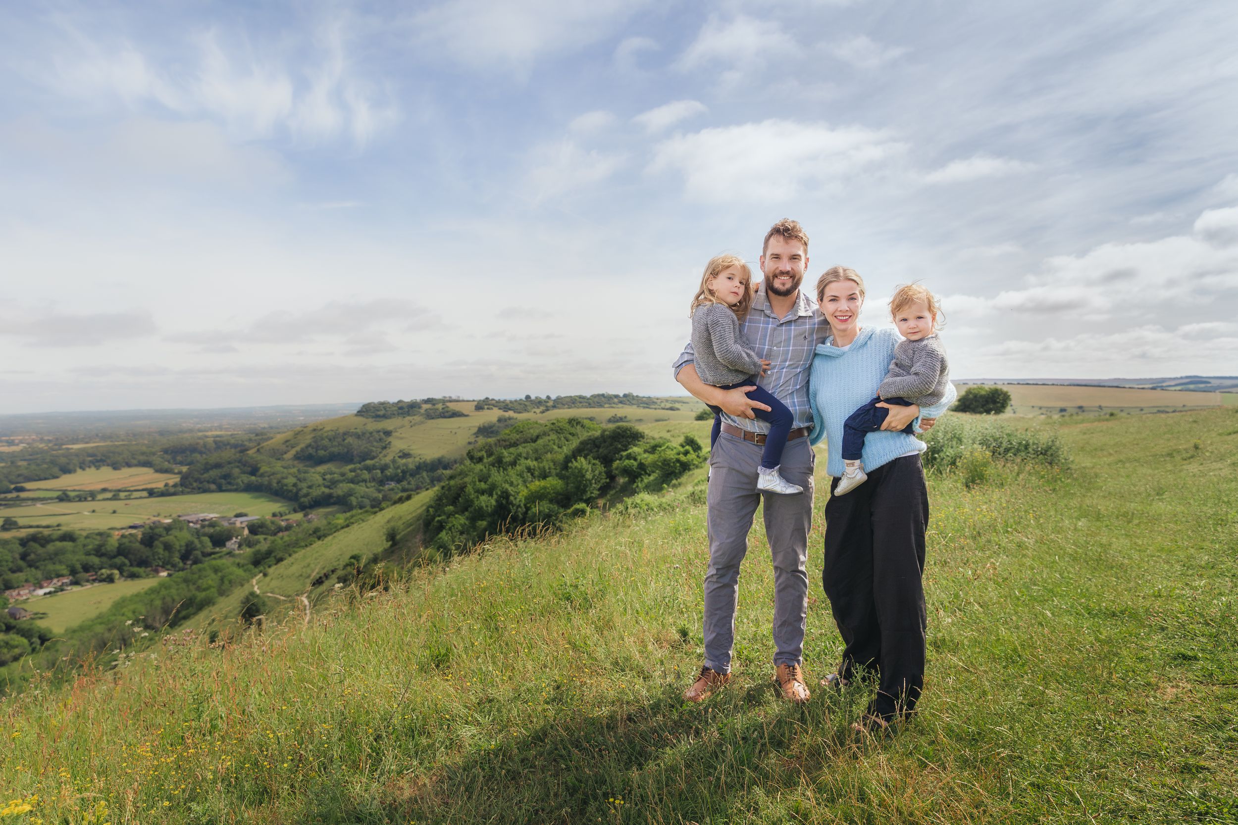 Family of four posing on a scenic green hilltop with rolling countryside views under a cloudy sky.