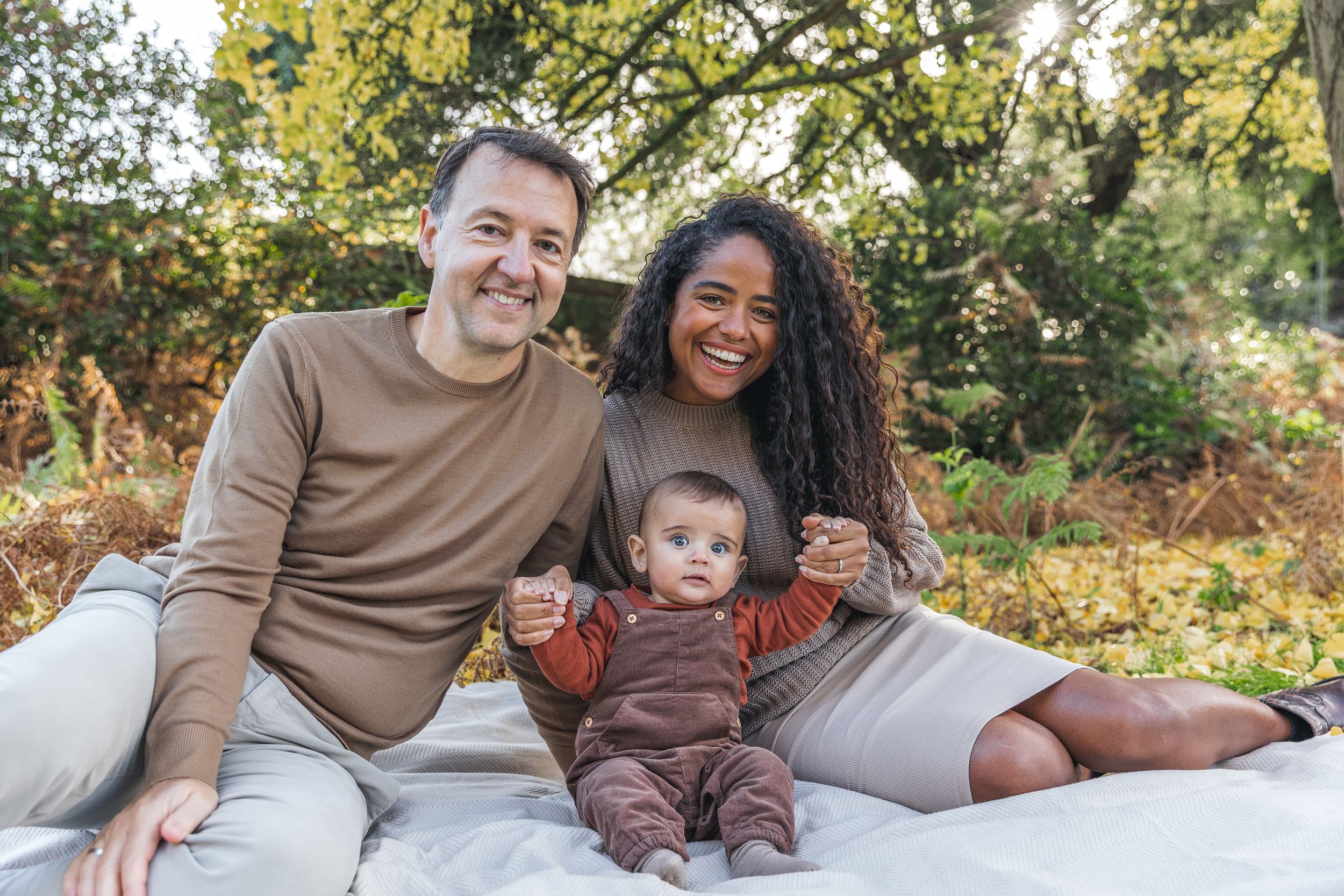 A happy interracial family with a baby sits on a blanket in a lush autumn park.