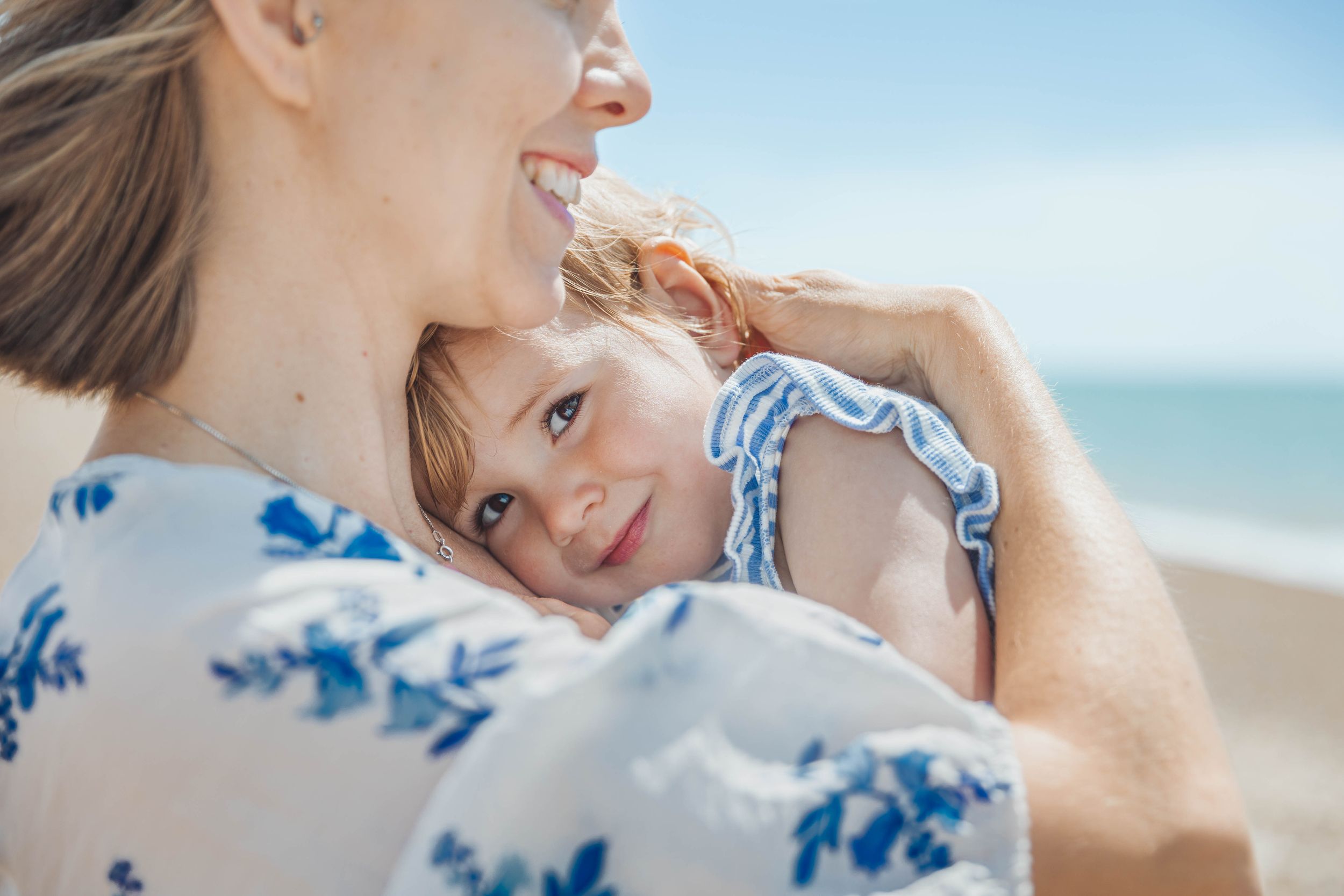A smiling mother holds her young daughter close at the beach, both sharing a joyful moment.