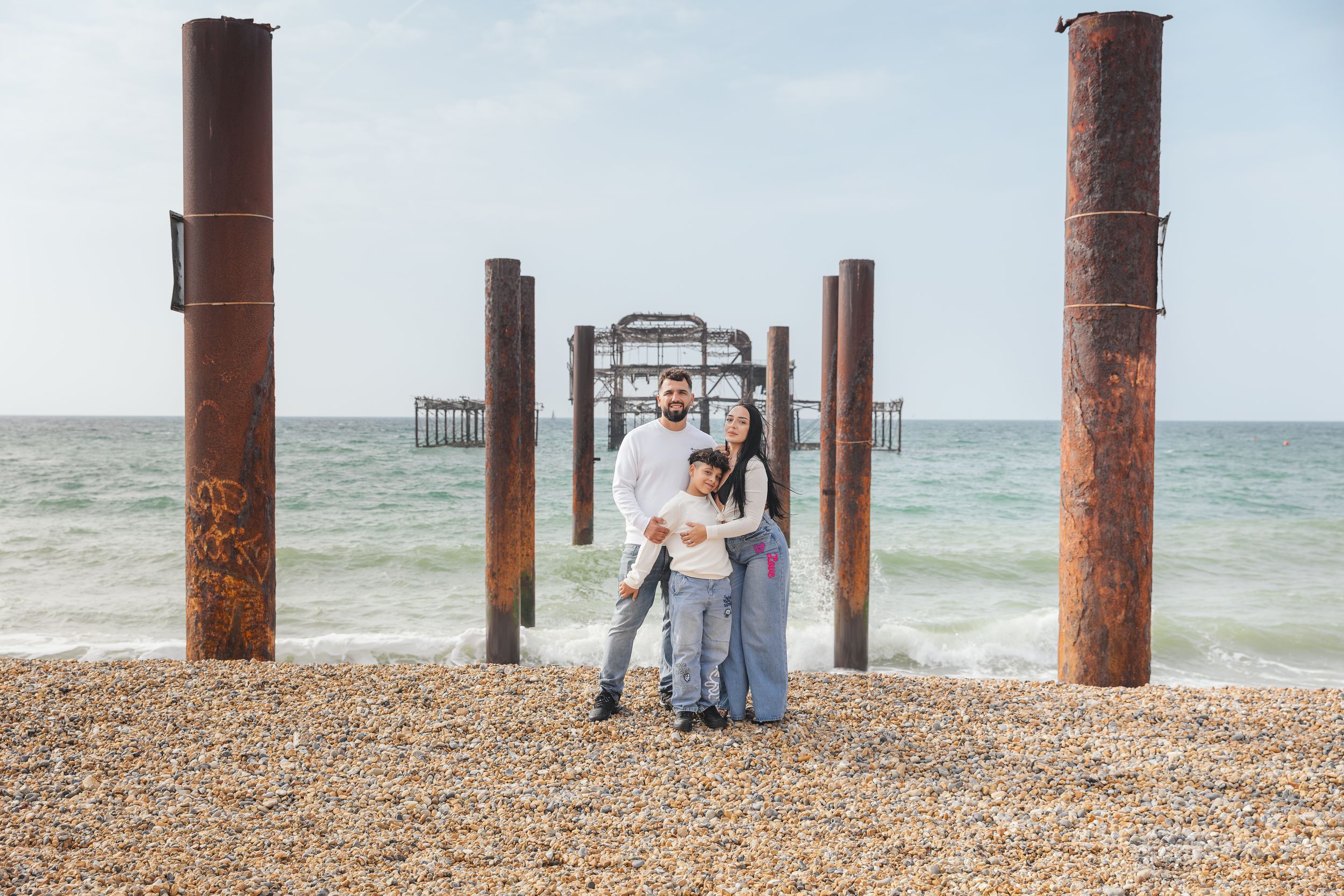 Family of three posing on a pebble beach beside rustic iron pier ruins with ocean waves in the background.