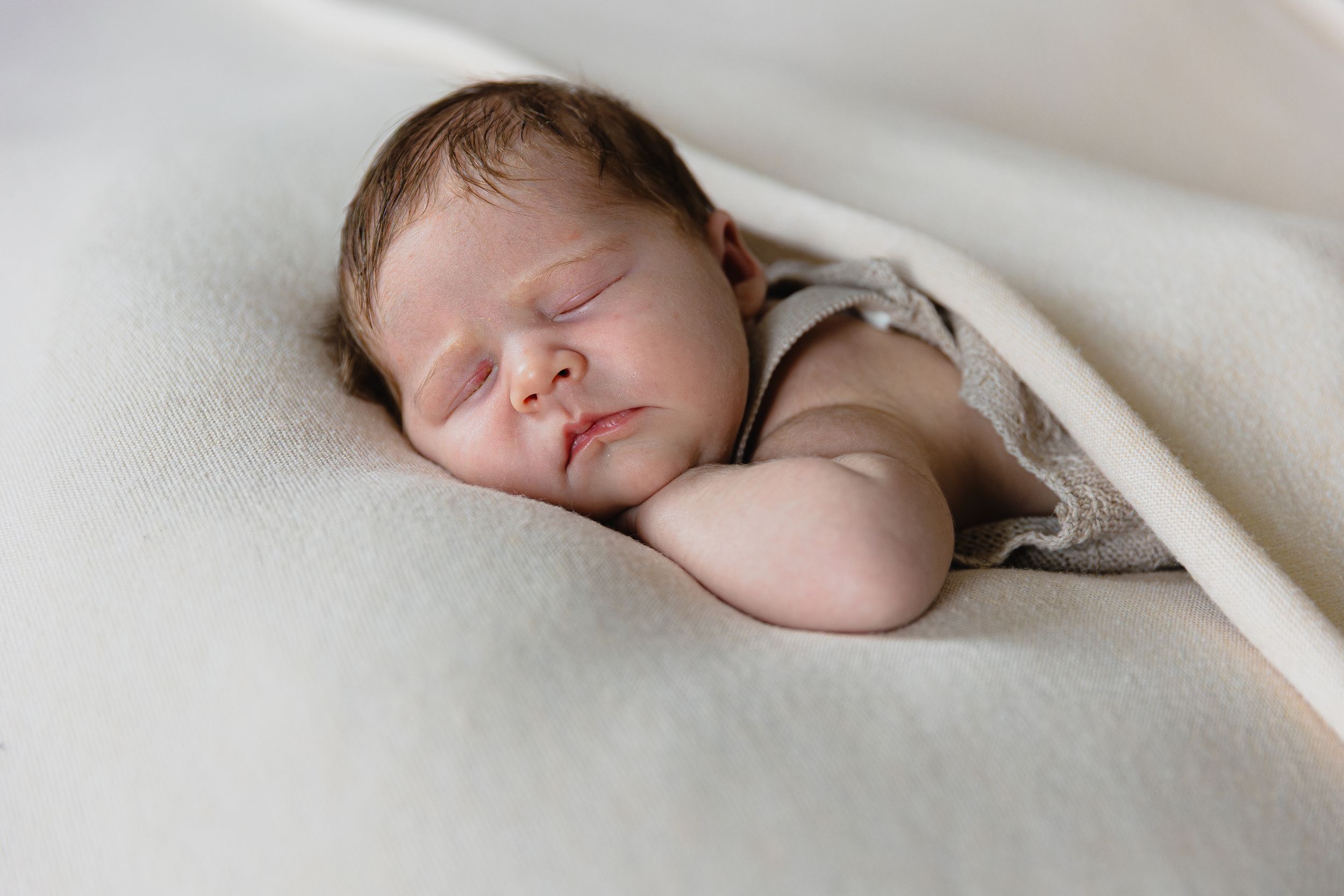 A peacefully sleeping newborn baby rests on a soft cream blanket in a cozy pose.