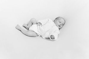 Black and white photo of a sleeping newborn baby in a white outfit on white background.