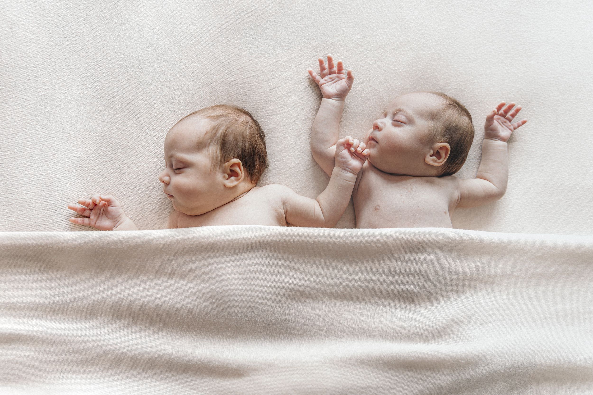 Two newborn twins lie side by side on a soft white blanket with tiny hands raised.