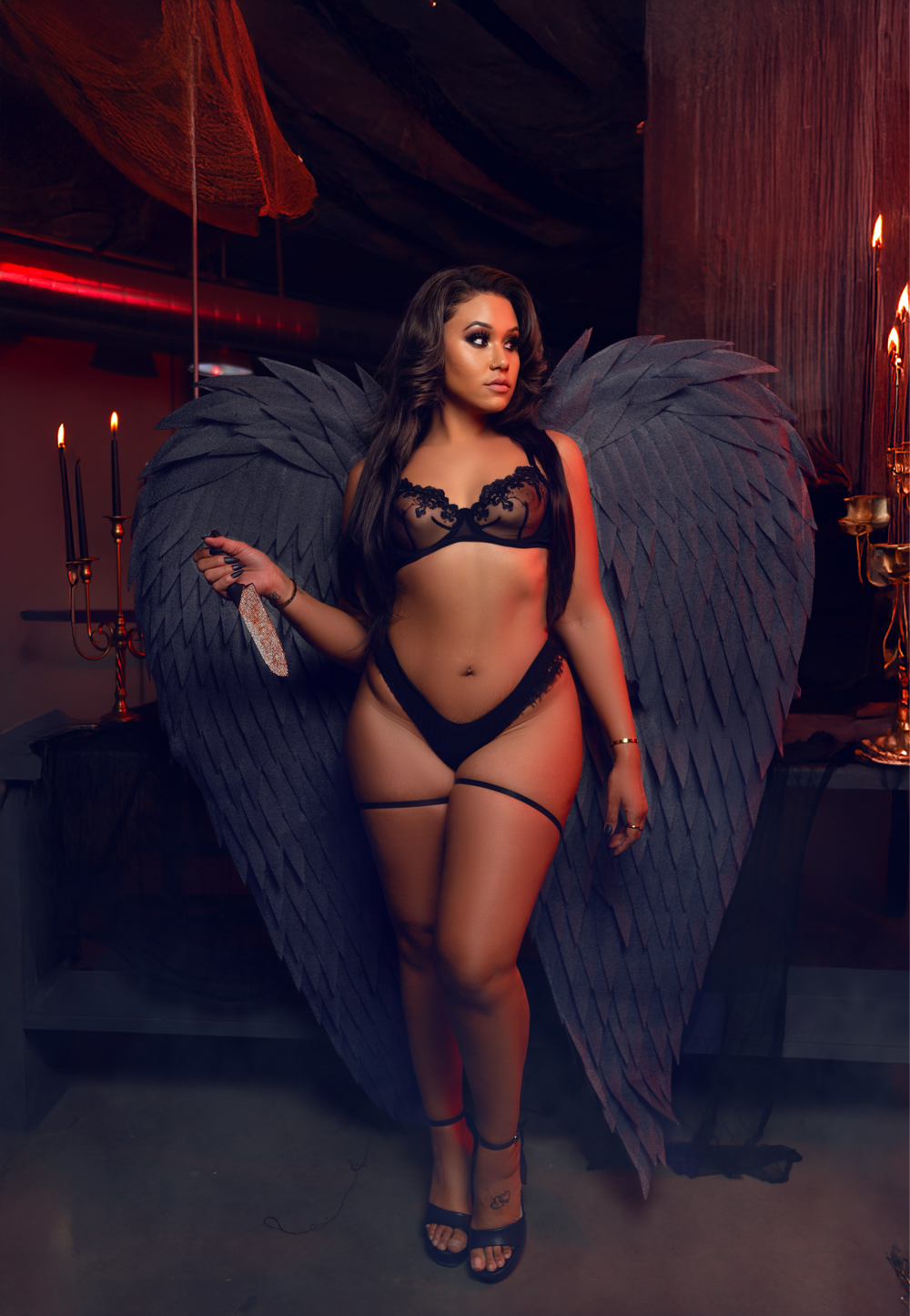 Dark angel figure in black lingerie and large wings poses dramatically in moody red-lit setting with candles.