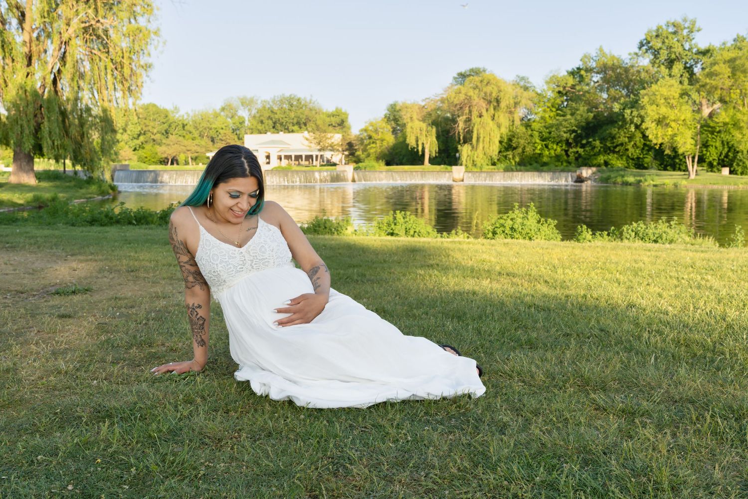 Expectant mother in white dress poses by scenic lake during golden hour.