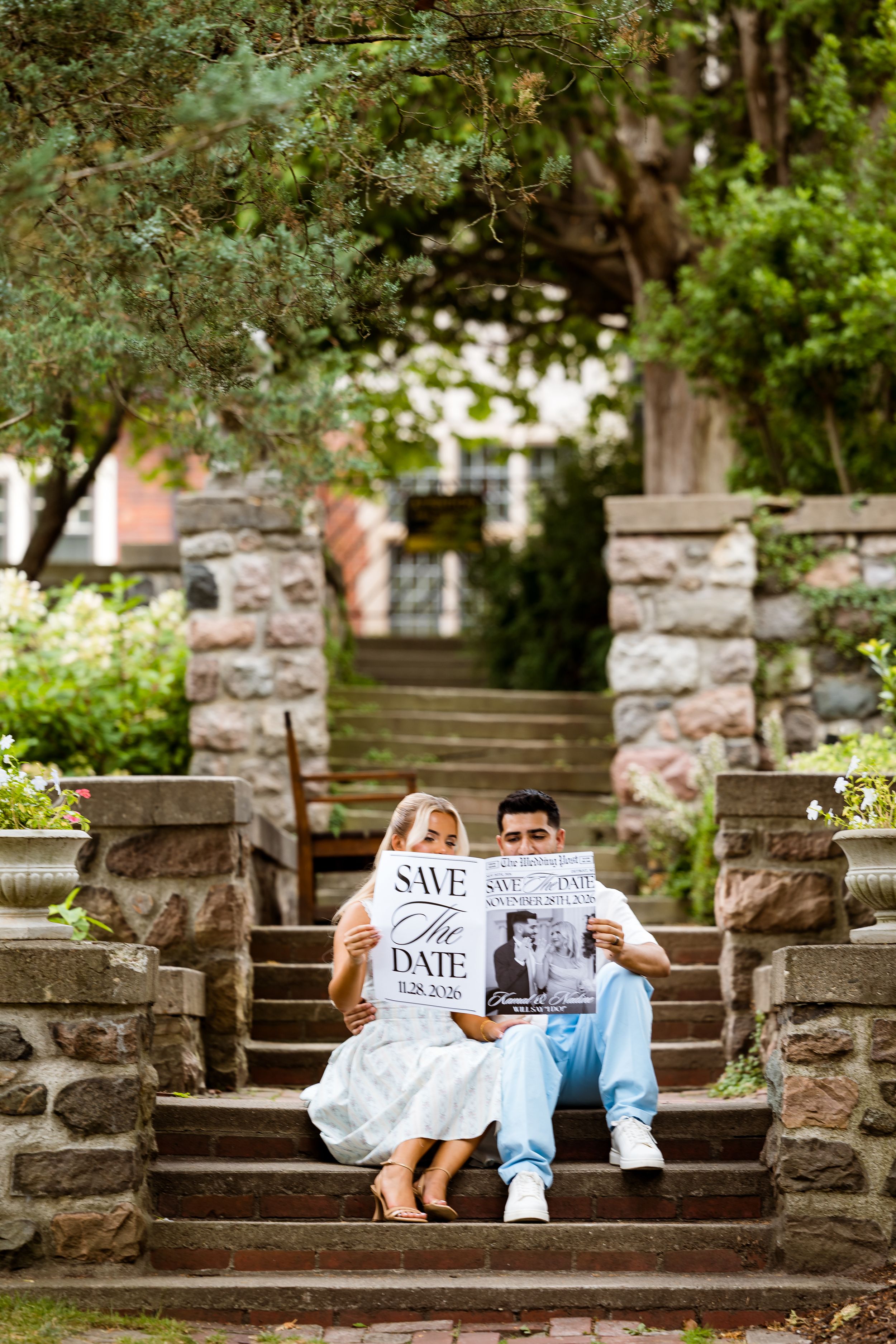 Couple sitting on stone steps holding a Save the Date sign surrounded by lush greenery and stone architecture.