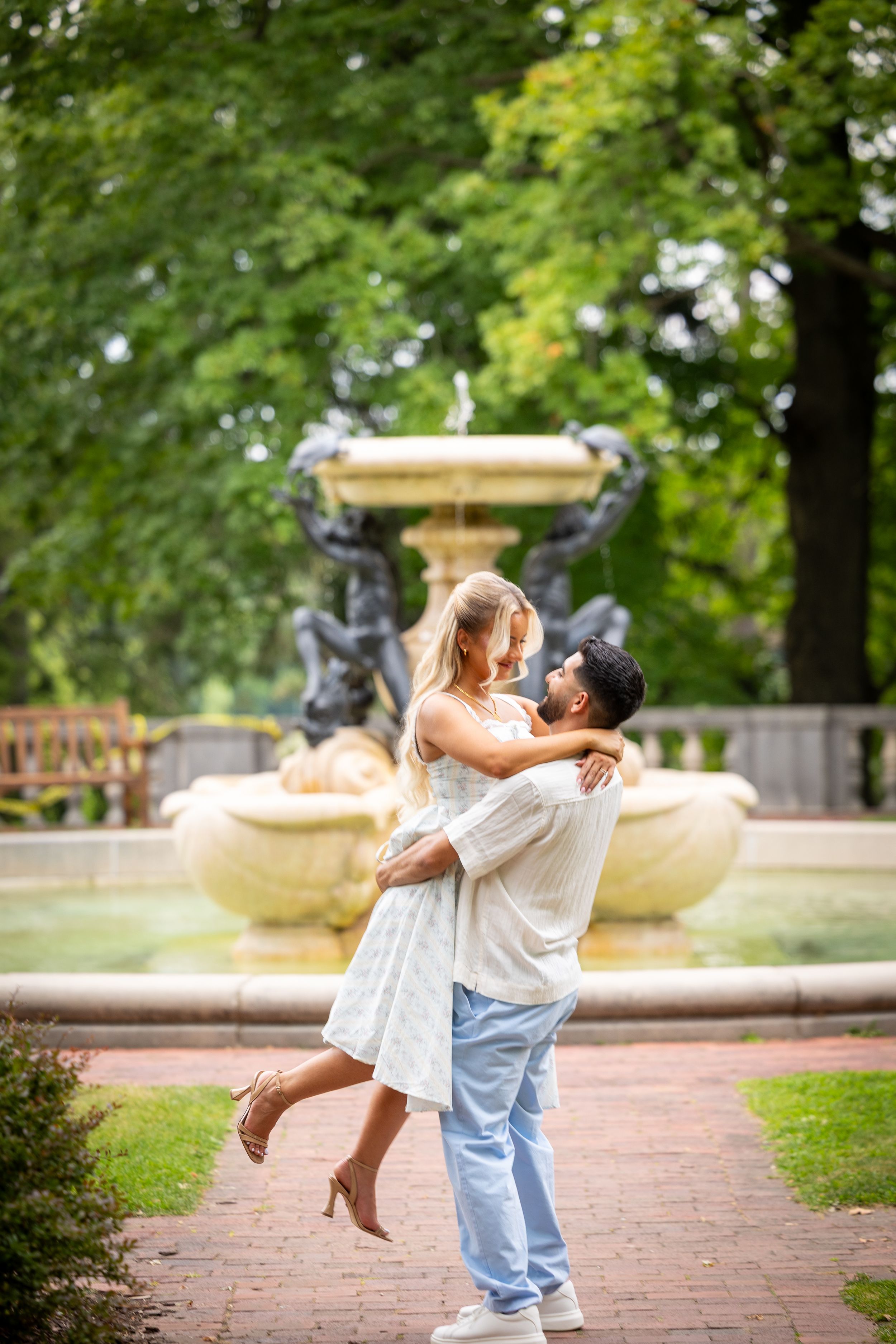 Couple embracing by ornate park fountain, man lifting woman in white dress, lush green trees background.