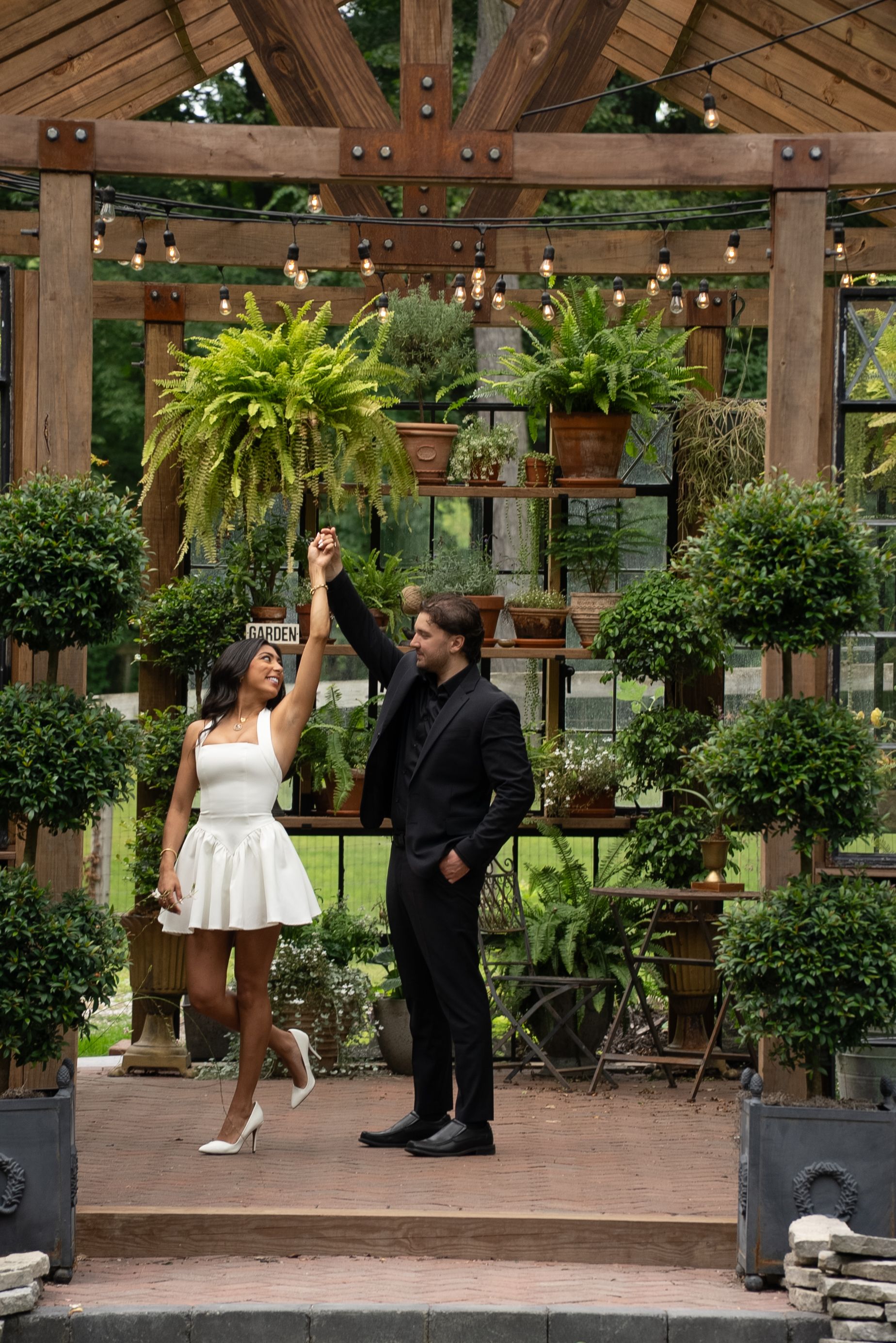 A couple stands together in a wooden pergola surrounded by lush greenery and potted plants in an outdoor garden setting.