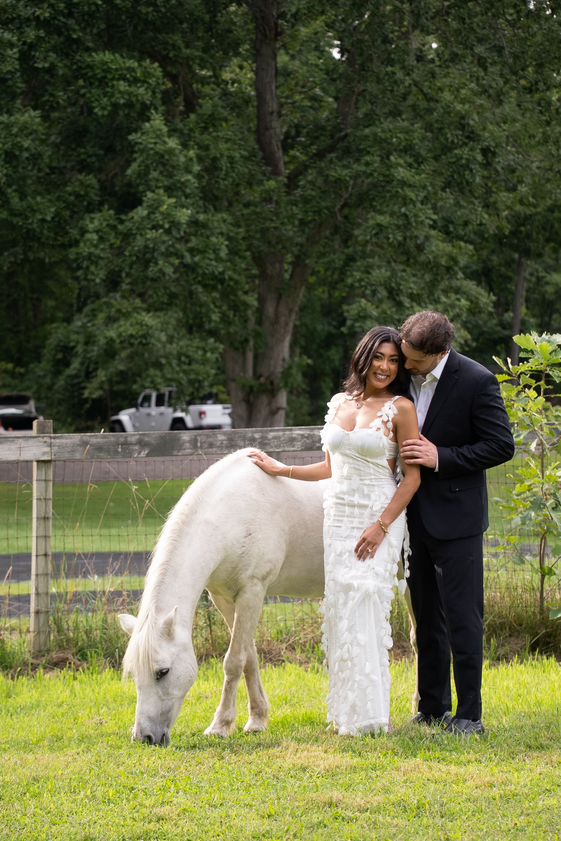 A couple in wedding attire stands in a grassy field with a white pony grazing beside them.