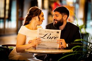 Couple smiling at each other while holding a newspaper with the word Love at a cafe table.