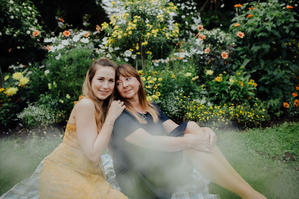 Two people in elegant dresses pose together in a lush garden setting with yellow flowers and greenery.
