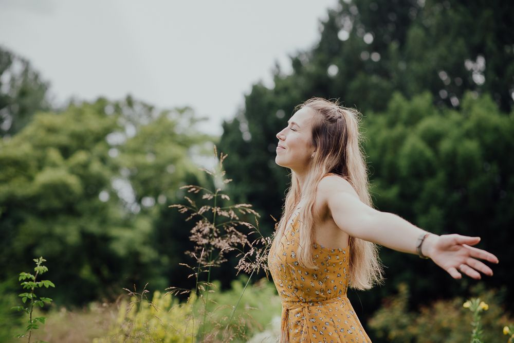 Eine Person in einem gelben Kleid steht mit ausgebreiteten Armen in einer üppigen Gartenlandschaft und drückt Freude und Freiheit aus.