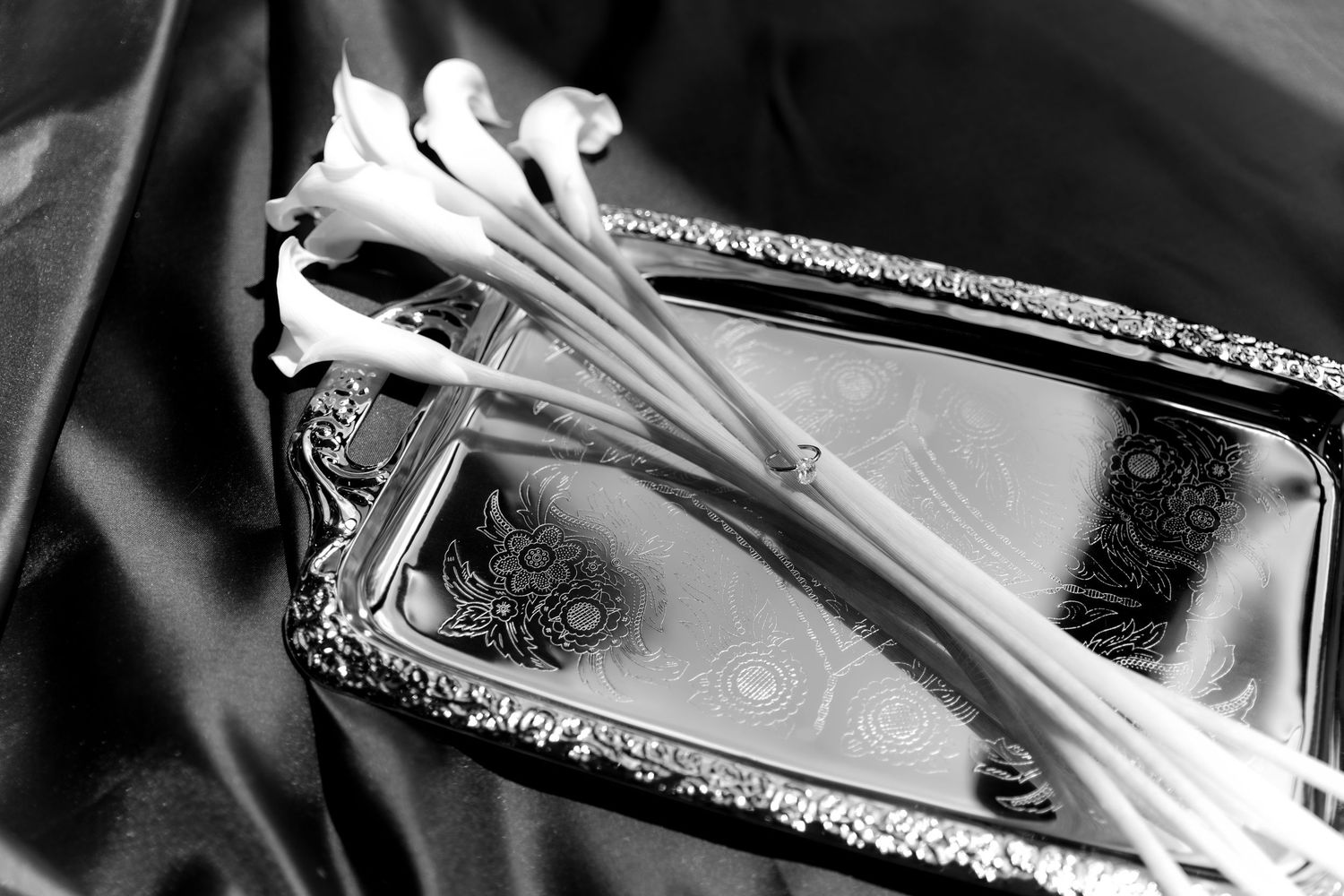 Black and white photo of white calla lilies resting on an ornate silver tray atop dark satin fabric.
