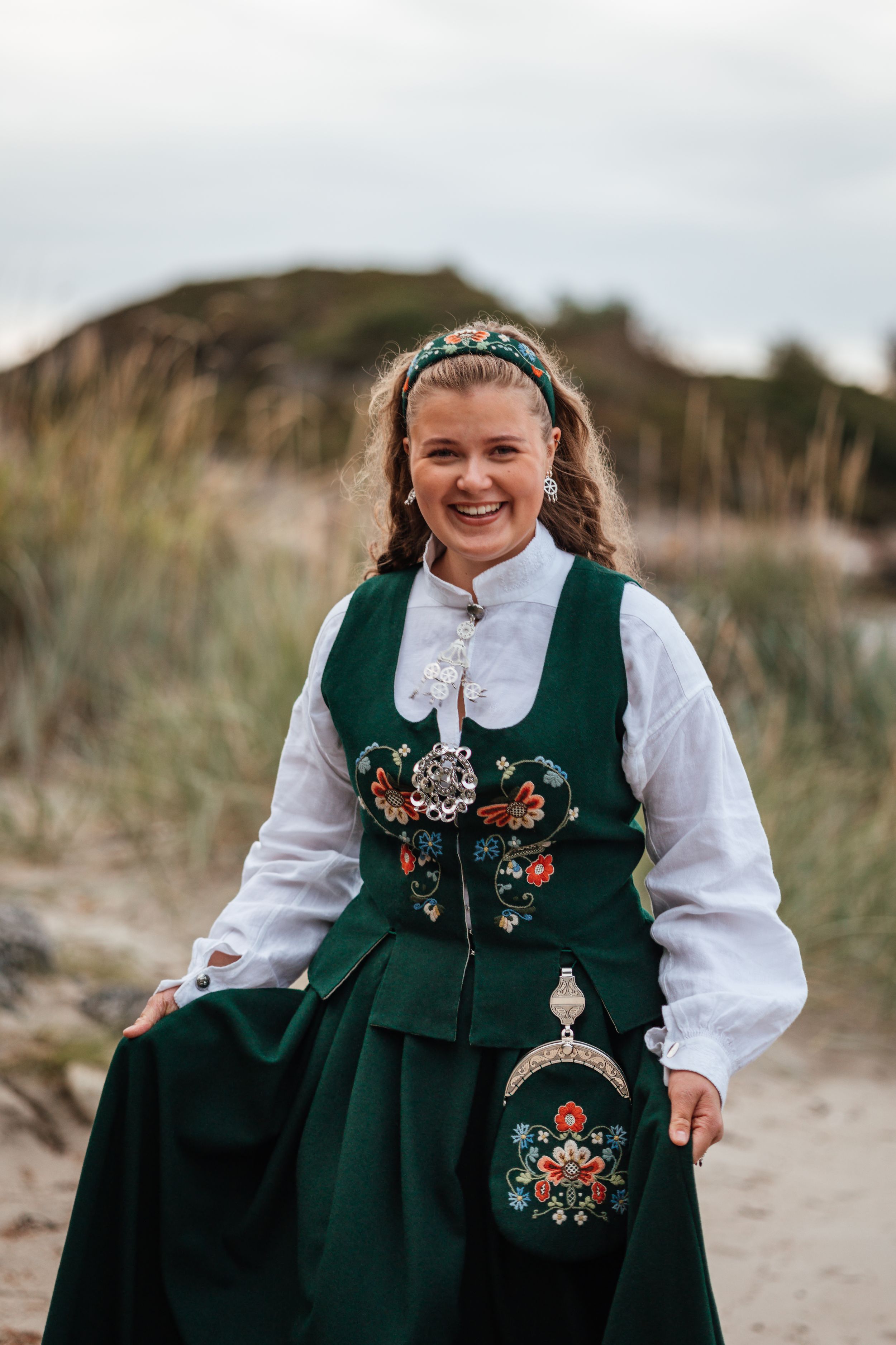 Smiling young woman wearing traditional Norwegian bunad dress in green with floral embroidery outdoors.