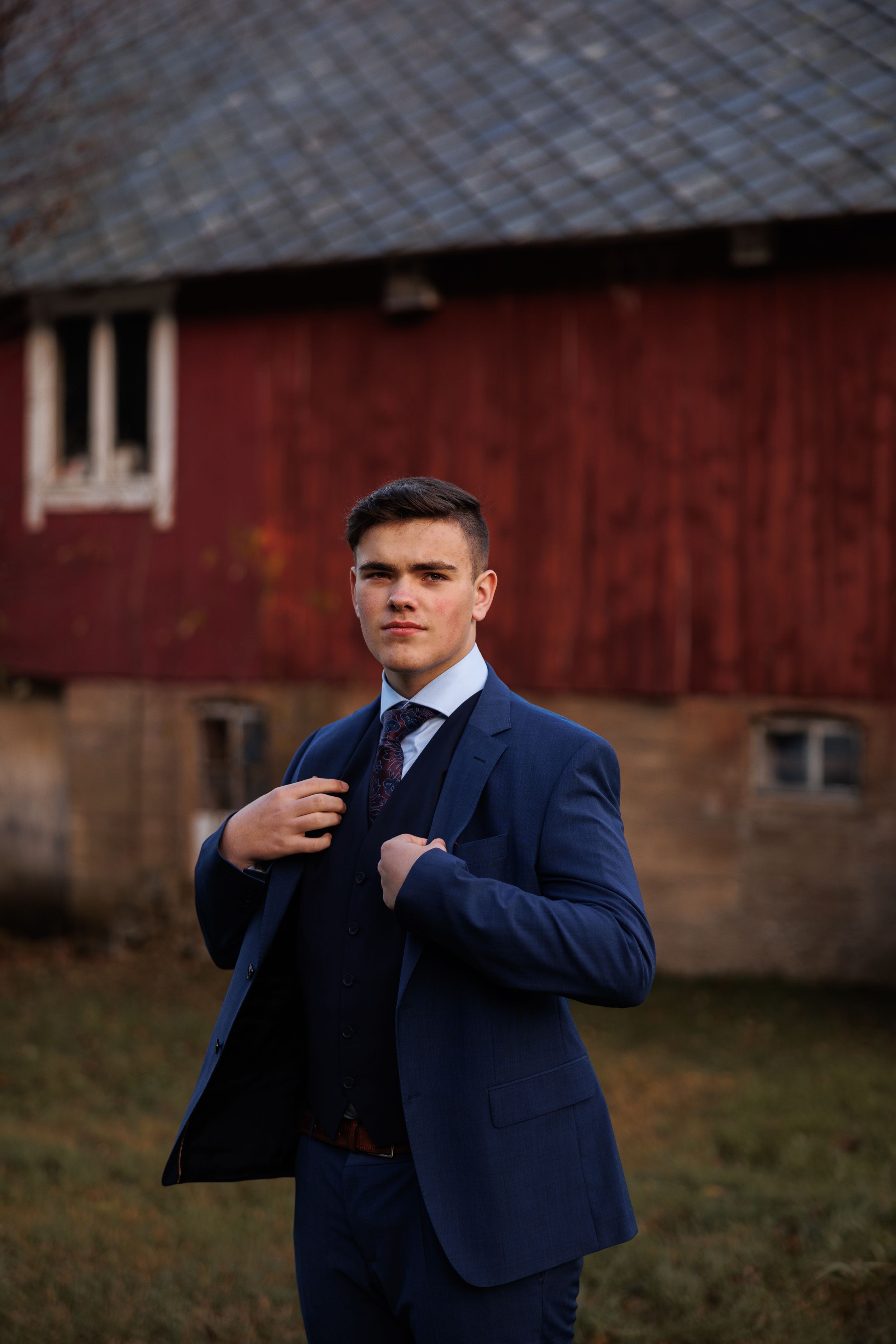 A formal portrait against a rustic red barn showcasing a navy blue three-piece suit.