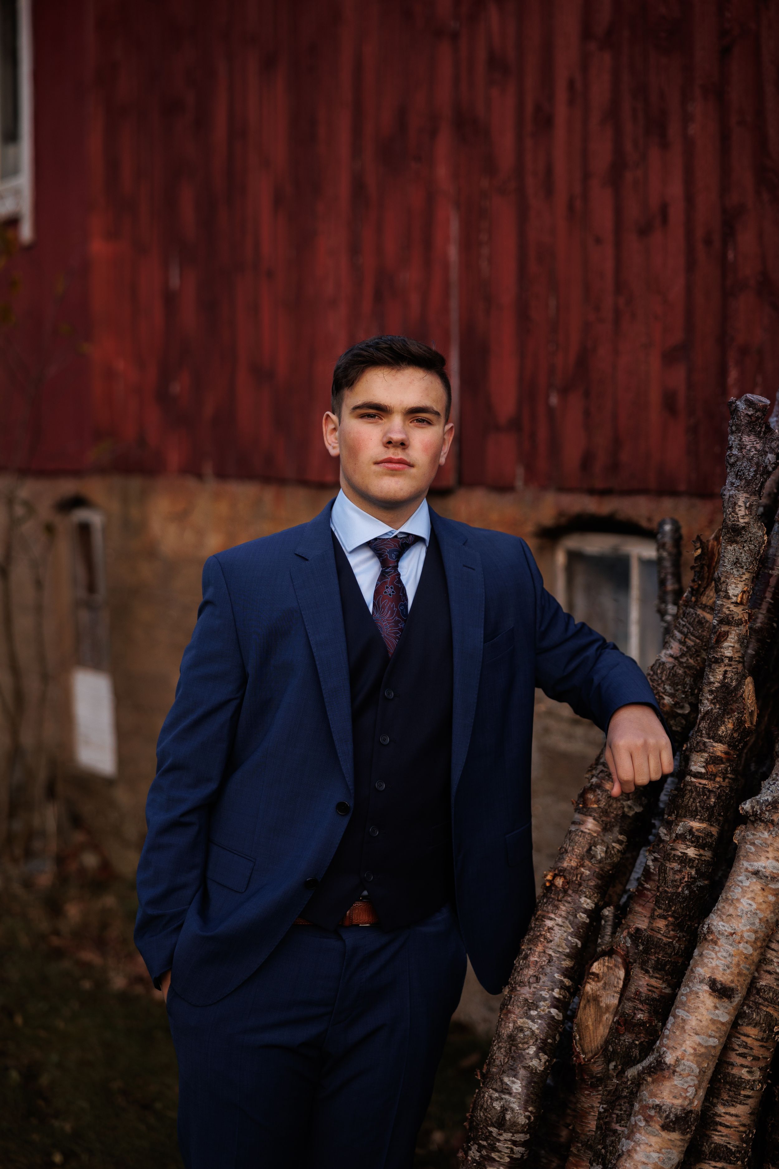 Navy suited figure poses confidently against weathered red barn siding and stacked firewood.