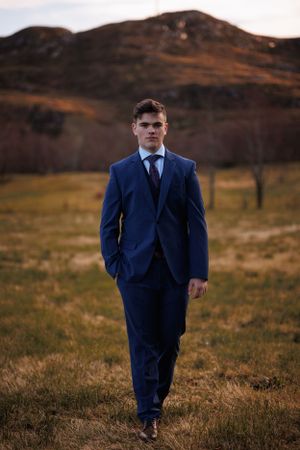 Professional portrait in a navy suit walking across Scottish highlands with mountains in background.
