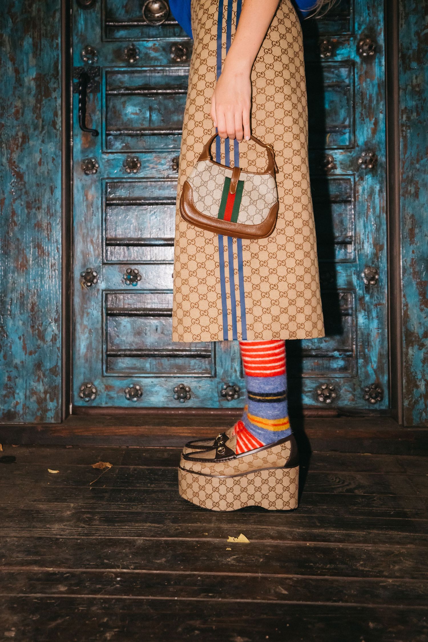 Gucci monogram skirt and colorful striped socks shown against weathered blue wooden doors.
