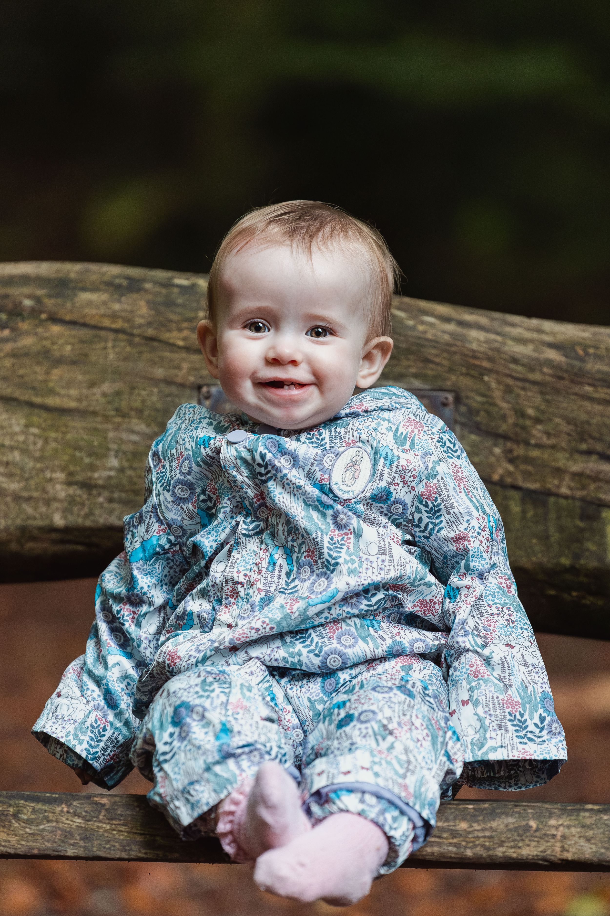 A small child in a patterned blue outfit sits on a wooden log outdoors.