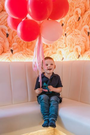 Young child sitting with red balloons against coral-colored backdrop.