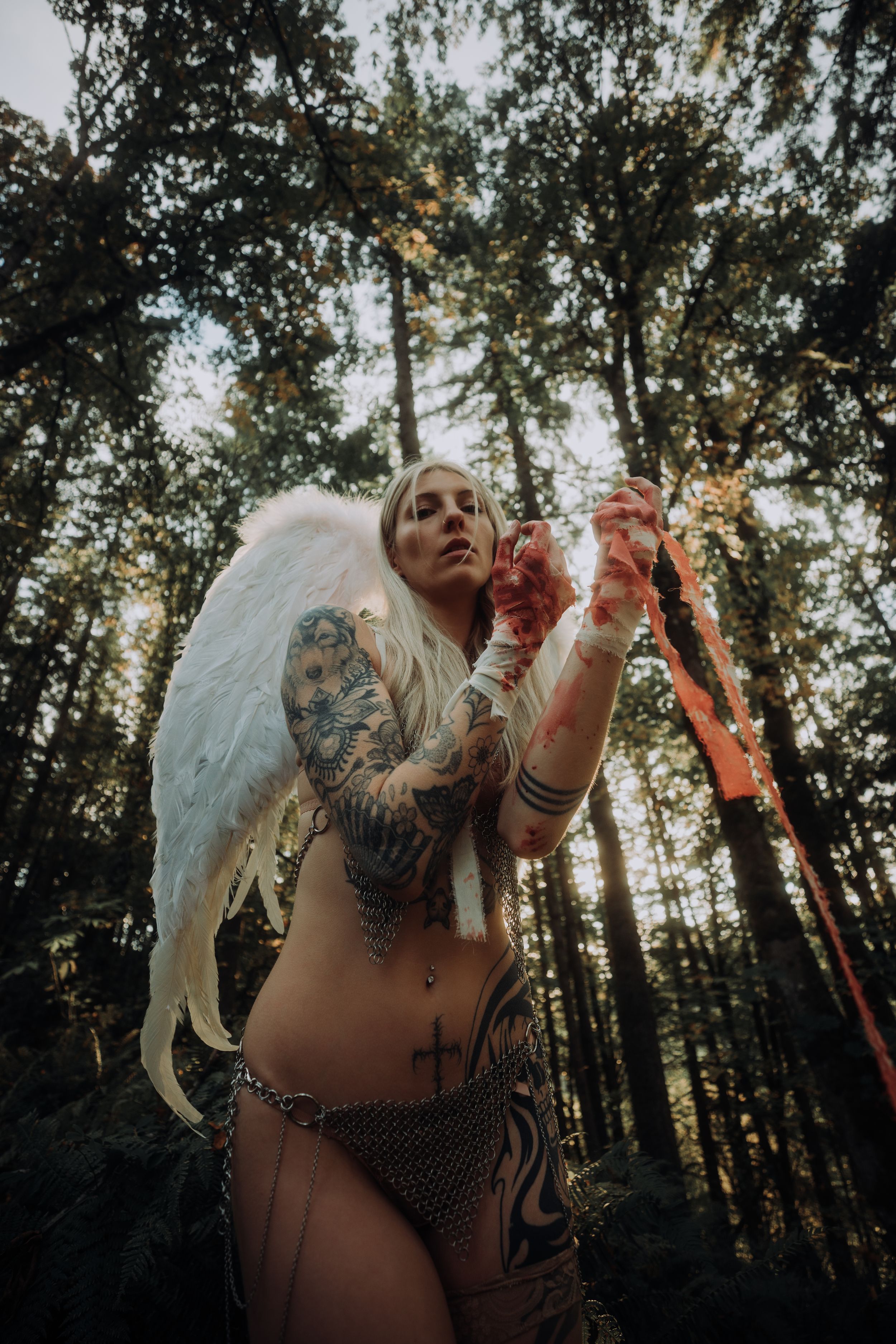 Artistic forest photoshoot featuring a tattooed figure in black lingerie posing against sunlit trees at golden hour.