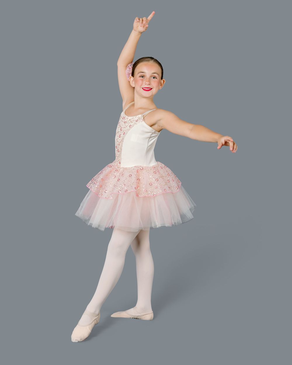 Young ballet dancer in pink tutu and white tights strikes graceful pose against gray background.