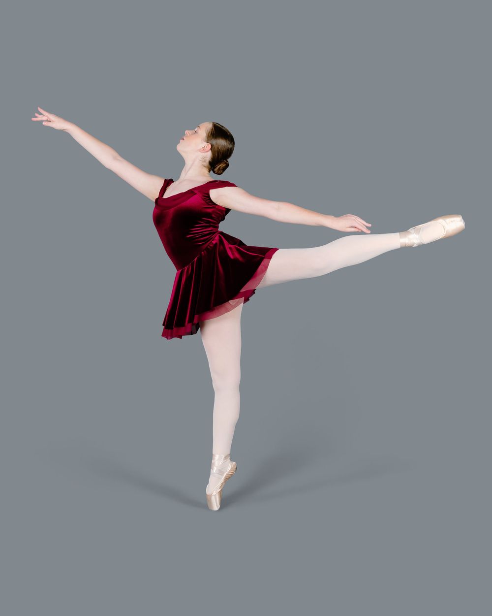A ballet dancer in white tights and burgundy leotard performs a graceful split leg turn in an arabesque position.