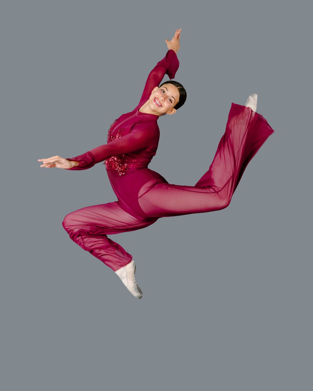 Dancer in burgundy jumpsuit leaps gracefully against gray background while performing contemporary dance move.