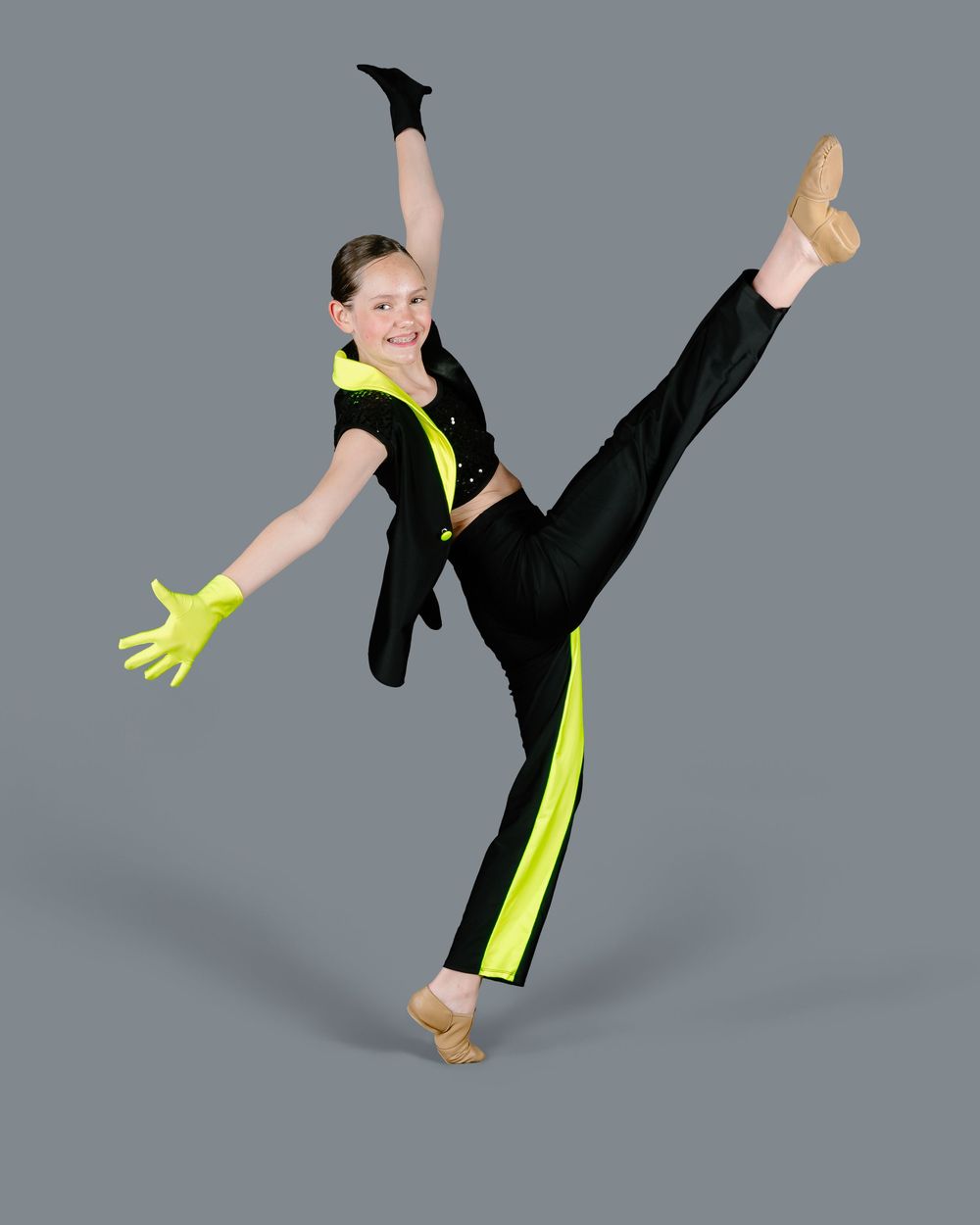 Dancer in black and neon yellow performance costume strikes dramatic high kick pose against gray background.
