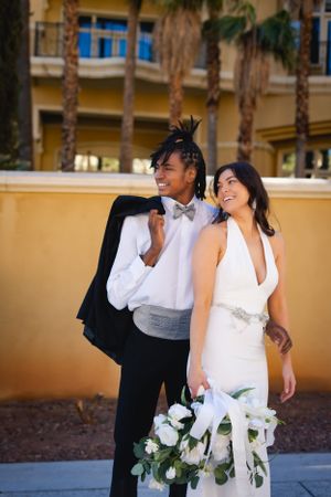 Stylish wedding couple smiling together outdoors with palm trees and white floral bouquet in a romantic pose.
