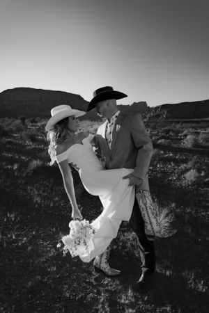 A romantic western-themed wedding photo shows a couple in cowboy hats embracing against a desert mountain landscape in black and white.