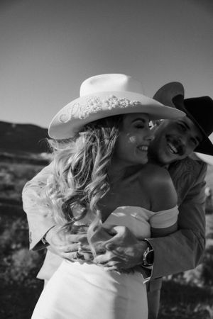 Black and white photo of a romantic couple embracing outdoors, wearing cowboy hats and western wedding attire.