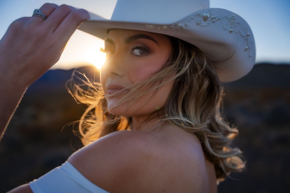 A white cowboy hat frames backlit figure at golden hour, with blonde hair gently moving in the breeze.