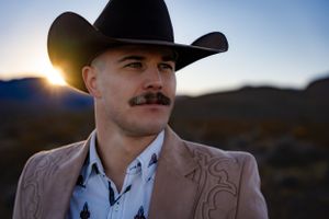 A person in a black cowboy hat and brown embroidered jacket poses at sunset in a desert landscape.
