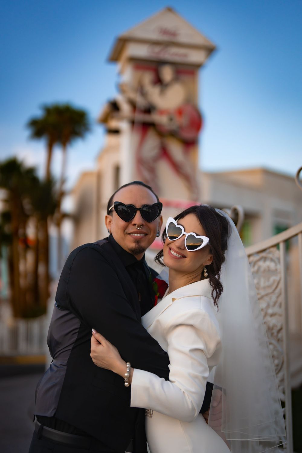 Newlyweds wearing sunglasses embrace and smile in front of the Little White Chapel at sunset.